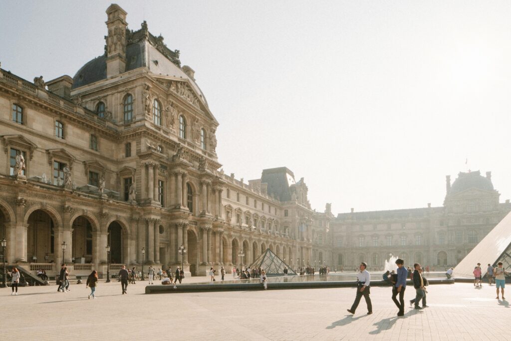 People walking at Louvre Museum courtyard with iconic pyramids in Paris during daytime.